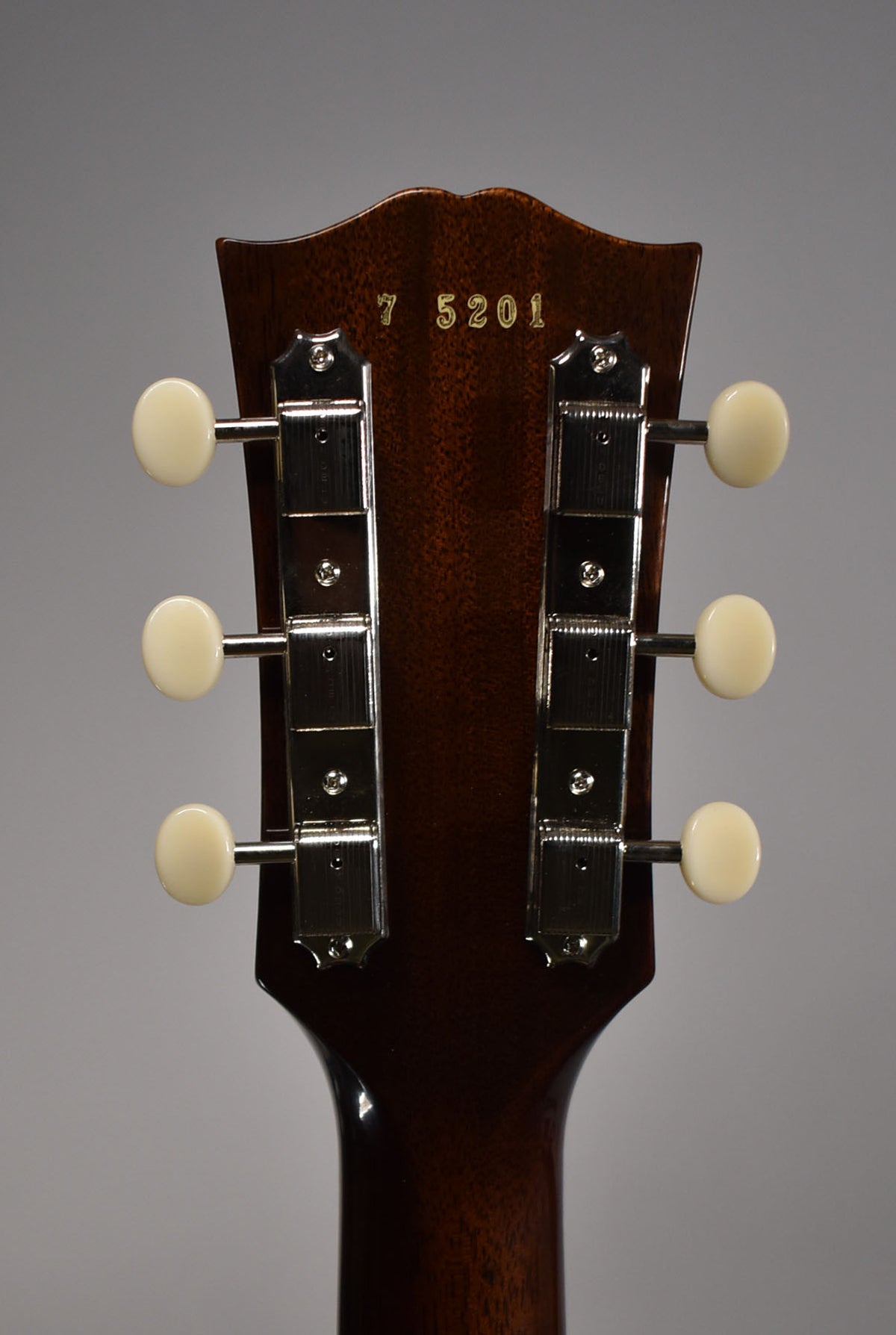 The guitar's dark brown headstock, viewed from behind, features six cream-colored tuning pegs and a gold serial number.