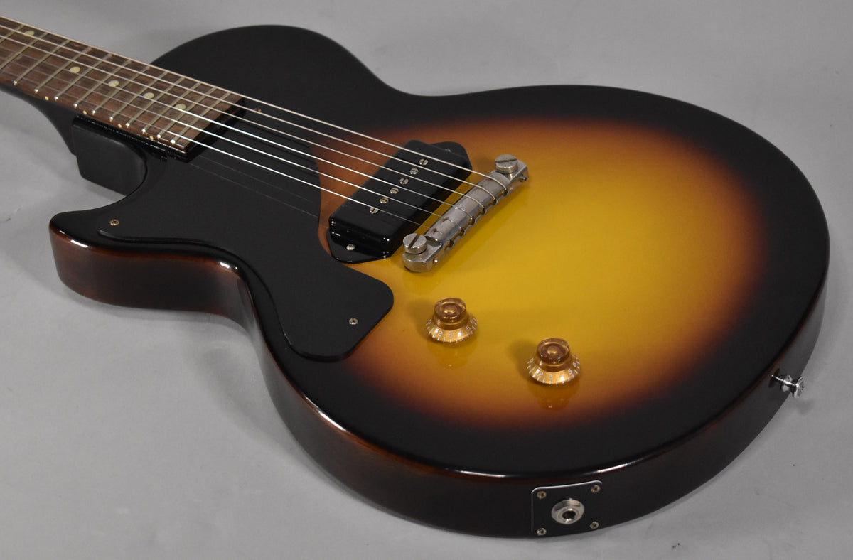 The sunburst single-cut guitar's body, with a black pickguard, P90 pickup, and two amber control knobs, is angled from the neck.