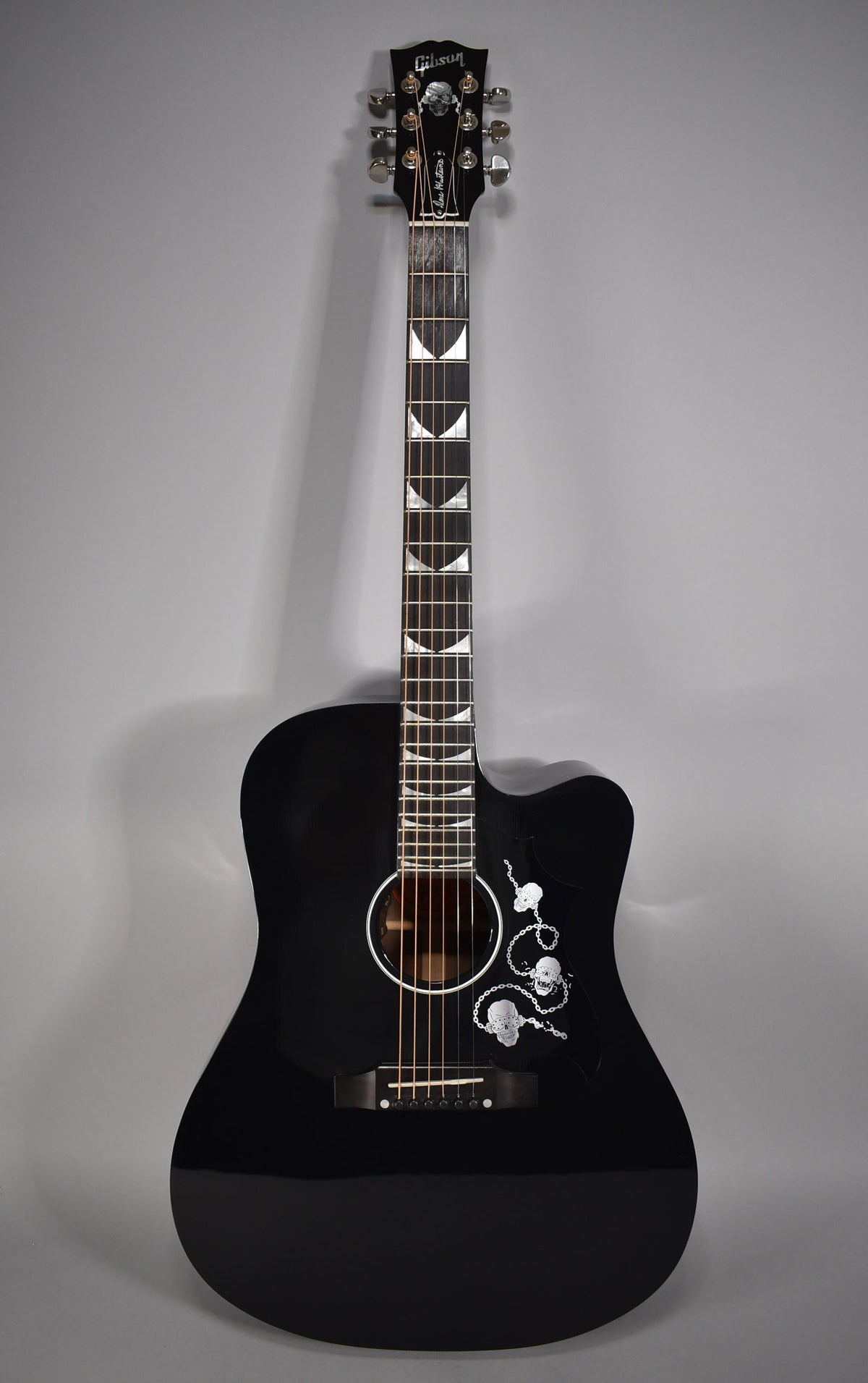 Full front view of a glossy black acoustic-electric guitar with a cutaway, featuring skull and chain inlay on the pickguard and crescent moon fret markers.