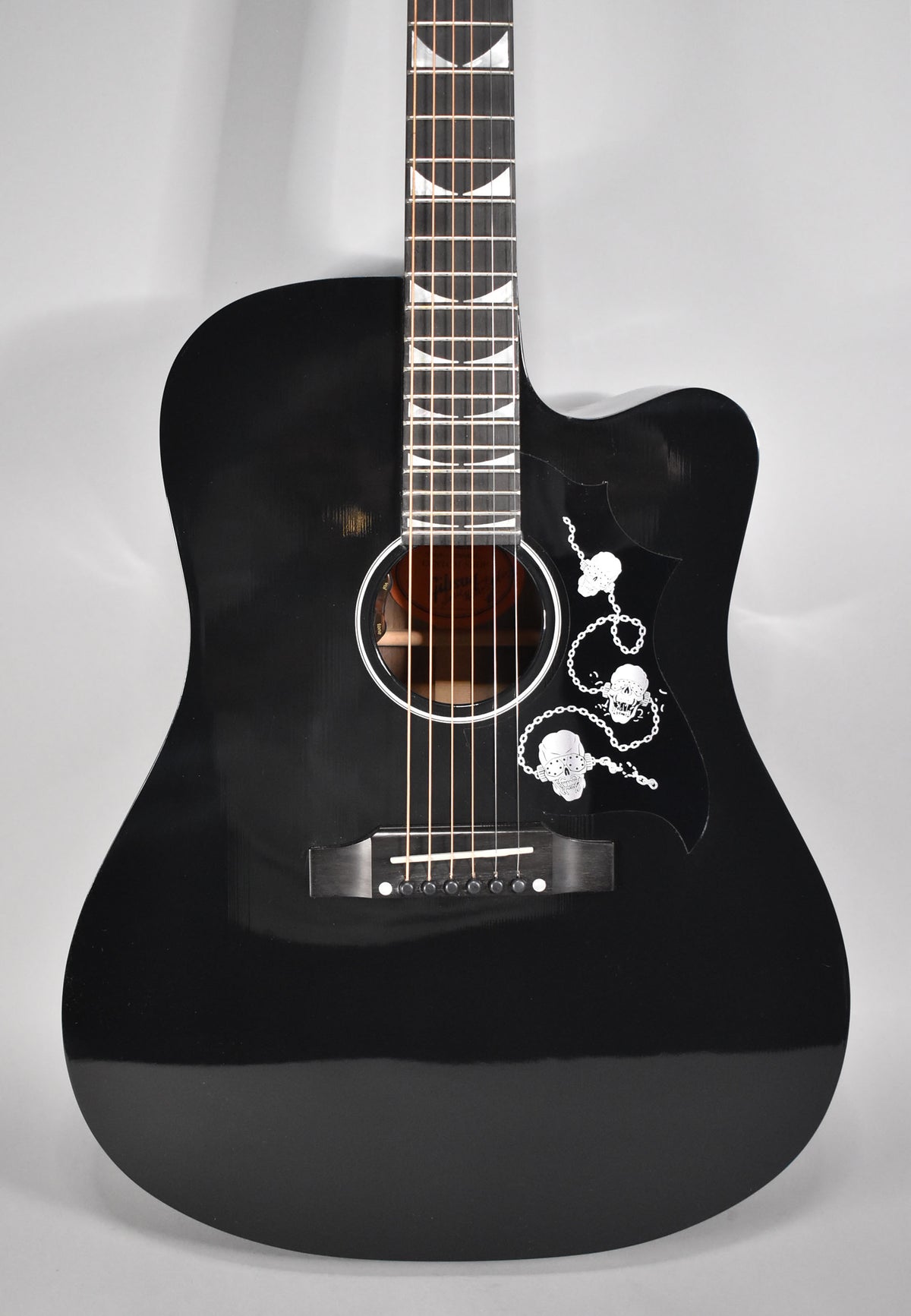 Full front view of a glossy black acoustic guitar with a cutaway, featuring a pickguard with white skull and chain graphics.
