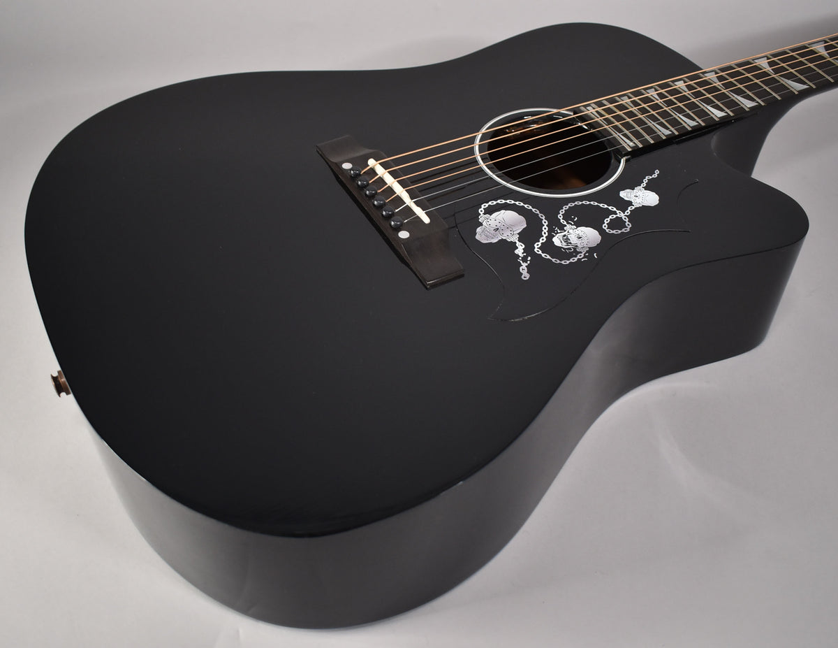 A black acoustic-electric guitar with a cutaway, skull and chain pickguard, and white fretboard inlays.