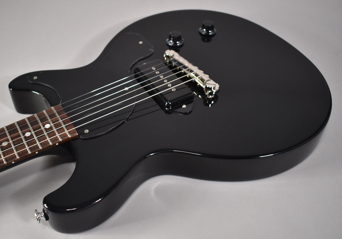 A glossy black double-cutaway electric guitar, angled from the lower bout, showing the P90 pickup, bridge, and two control knobs.