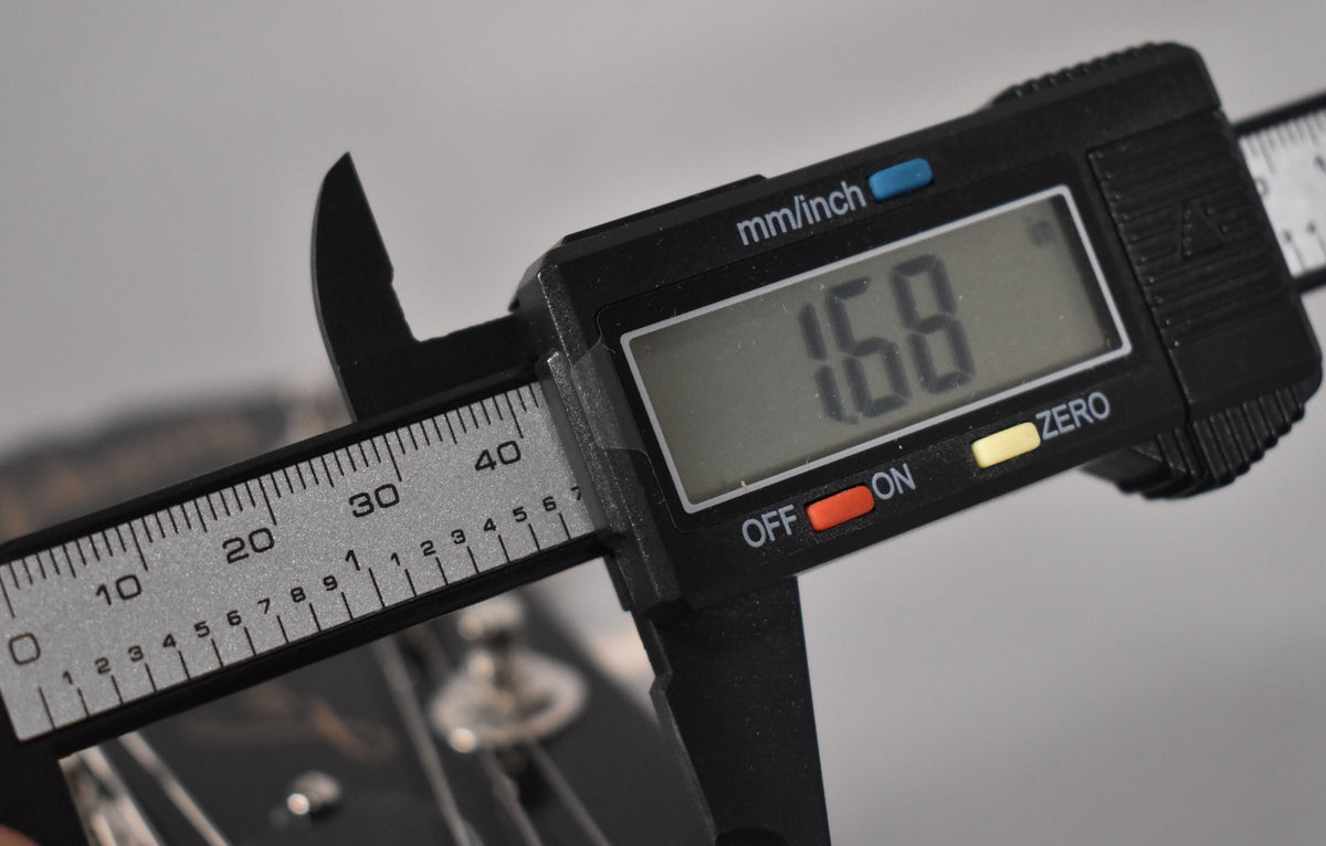 A digital caliper measures the guitar's nut width at 1.68 inches, with the headstock and tuning pegs blurred in the background.