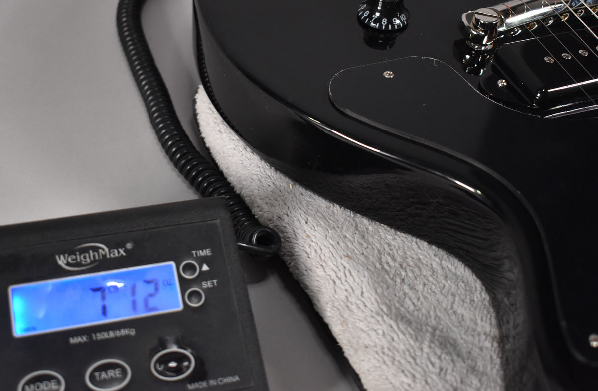 The guitar's glossy black body rests on a grey cloth, with a scale showing "7.12" lbs in the foreground.