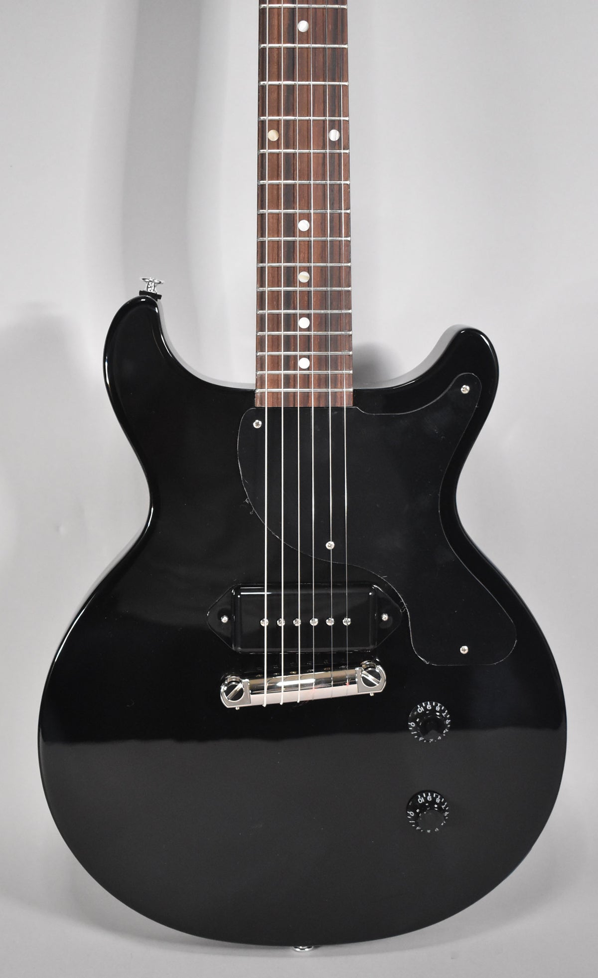 A full-frame, front view of a glossy black double-cutaway electric guitar with a single P90 pickup and two black control knobs.
