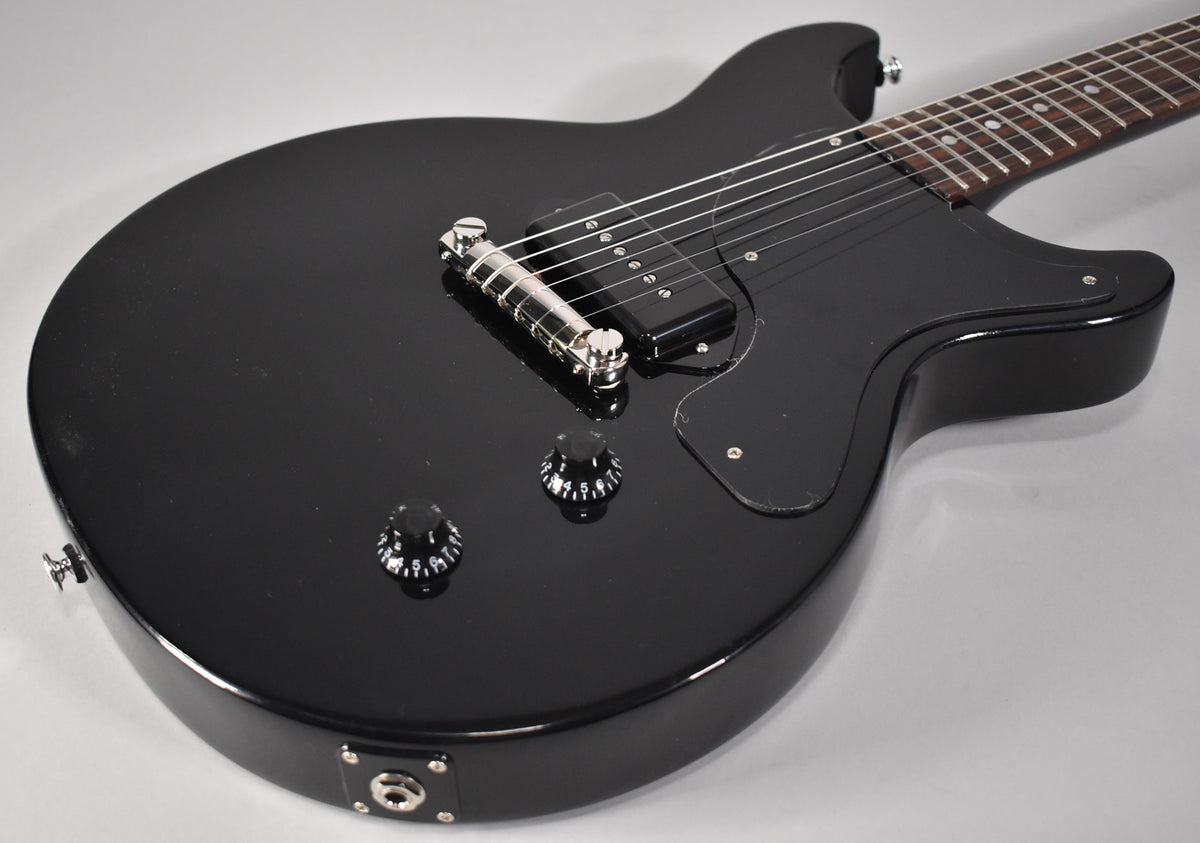 The glossy black double-cutaway electric guitar is angled from above, showing its single P90 pickup, chrome wraparound bridge, and two control knobs.