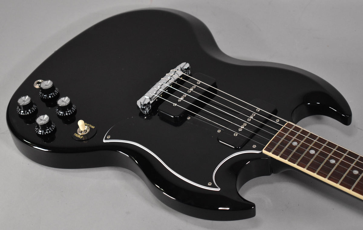 Glossy black SG body, angled from above, showing the lower bout, four black knobs, cream switch, black pickguard, and bridge.