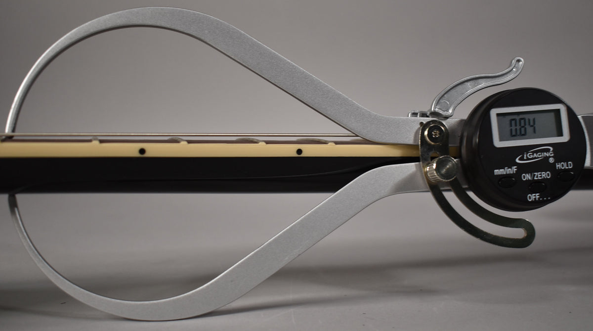 A digital caliper measures the guitar's ebony fretboard, showing a reading of 0.84, with cream binding and dot inlays.