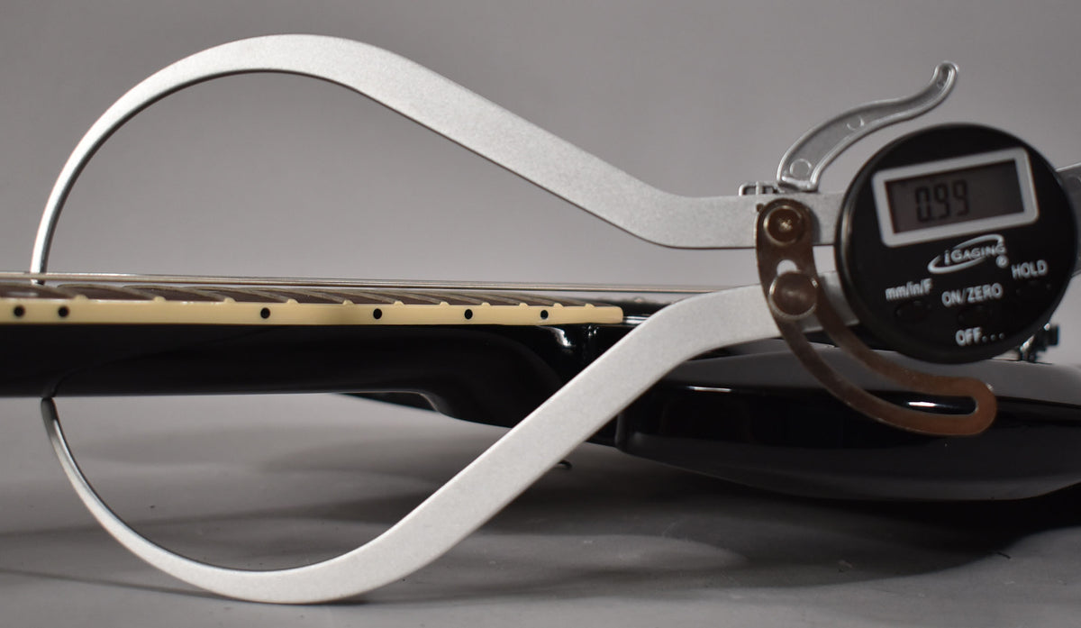 A digital string action gauge shows a 0.99mm reading on the ebony guitar's fretboard, with cream binding and dot inlays.