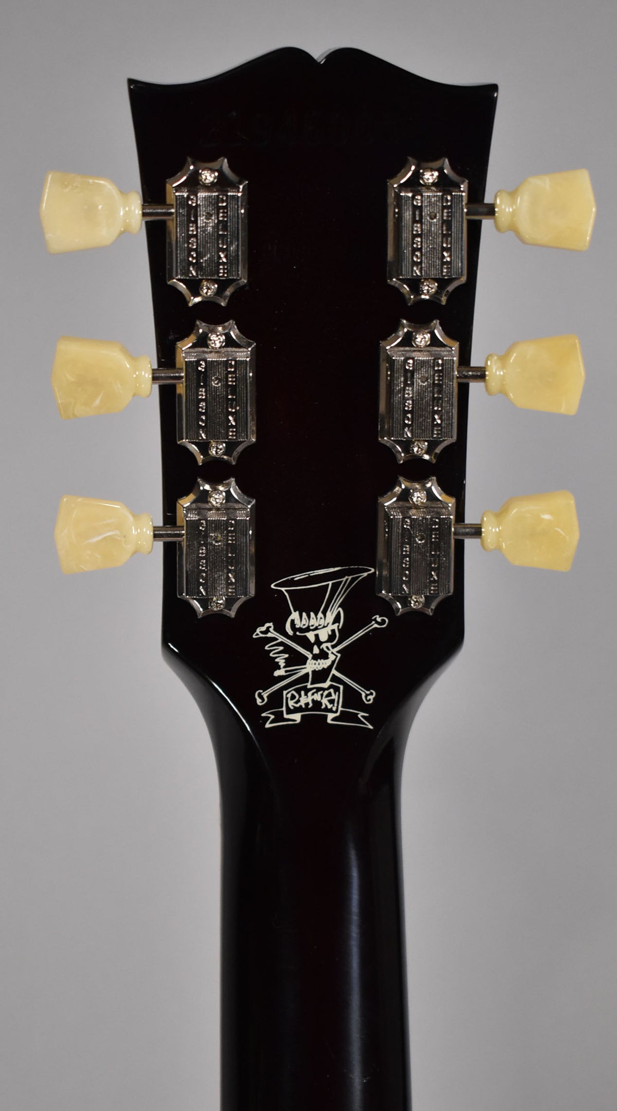 Rear view of the dark brown headstock and upper neck, showing six cream tuning pegs and a white skull and crossbones logo.