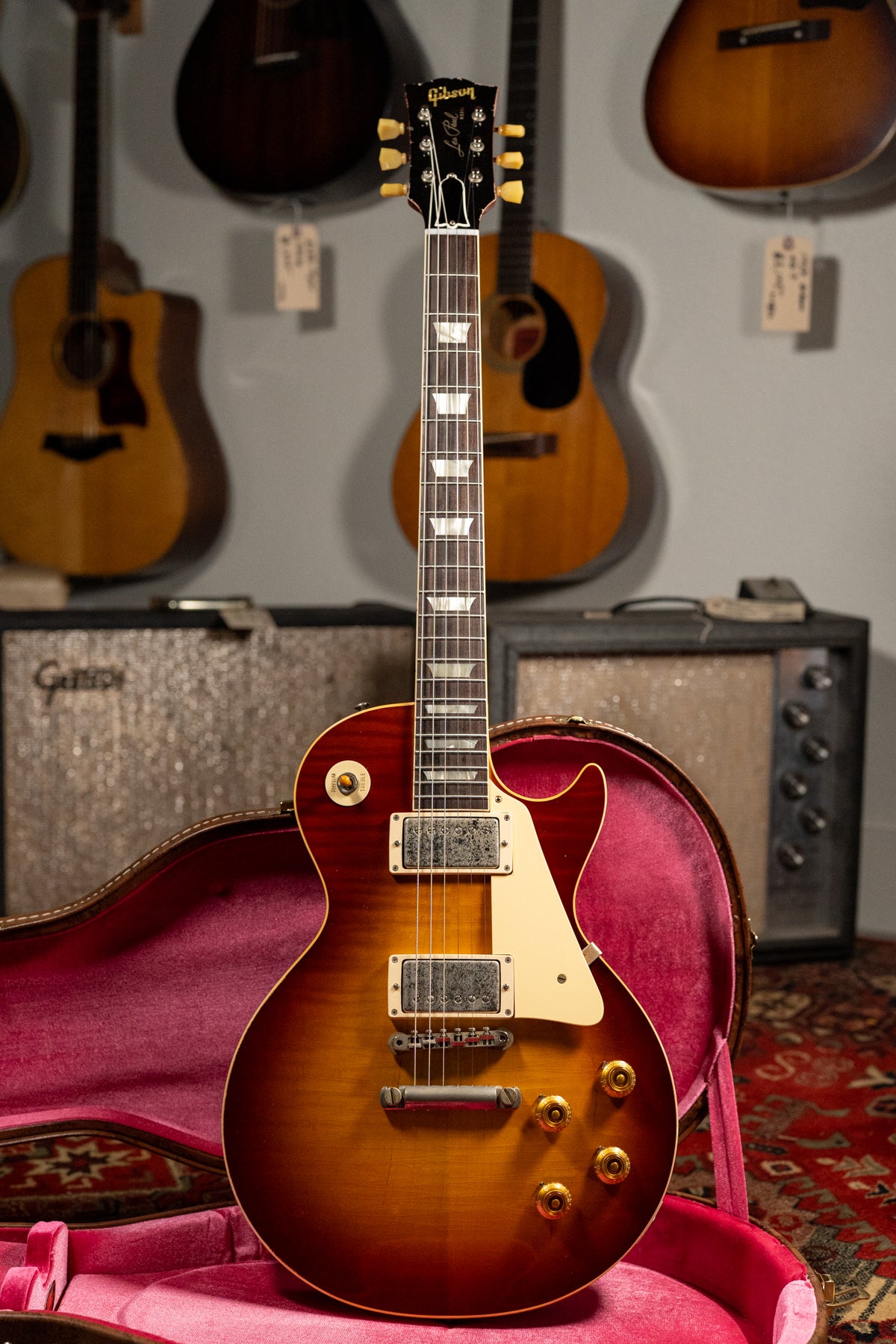 A full-length view of a sunburst electric guitar with aged hardware, resting in a red velvet-lined open hard case.