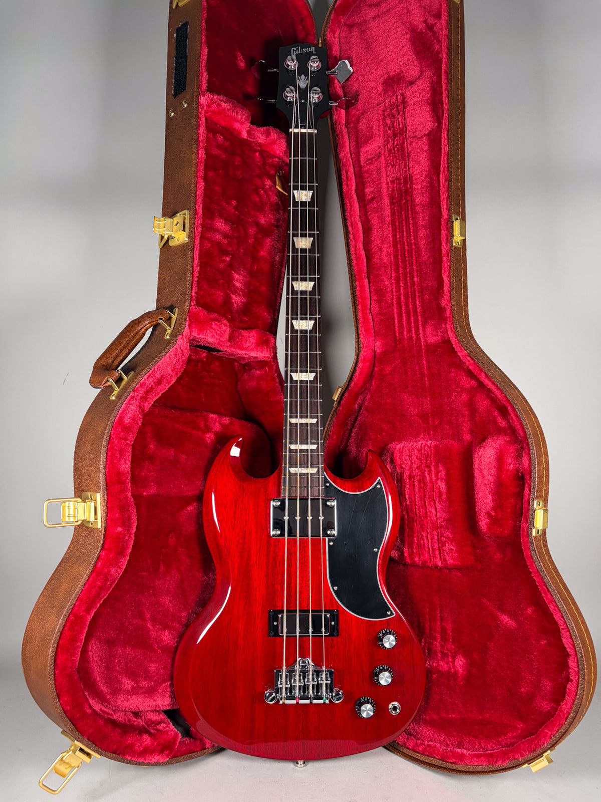 Full-length view of a glossy cherry red SG bass with a black pickguard, chrome hardware, and four control knobs, standing in its open brown case with red plush lining.