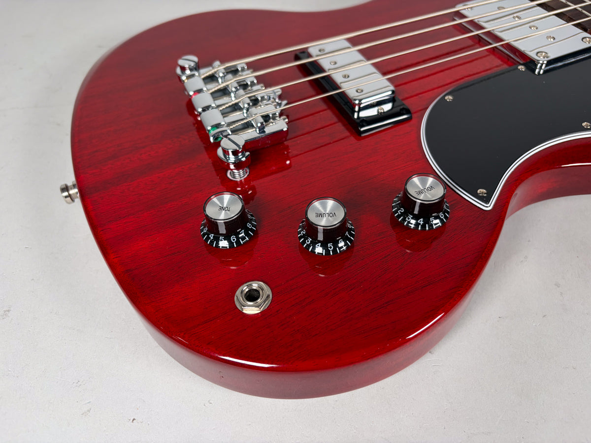 Close-up of the cherry red bass body, showing the chrome bridge, two pickups, black pickguard, three control knobs, and input jack.