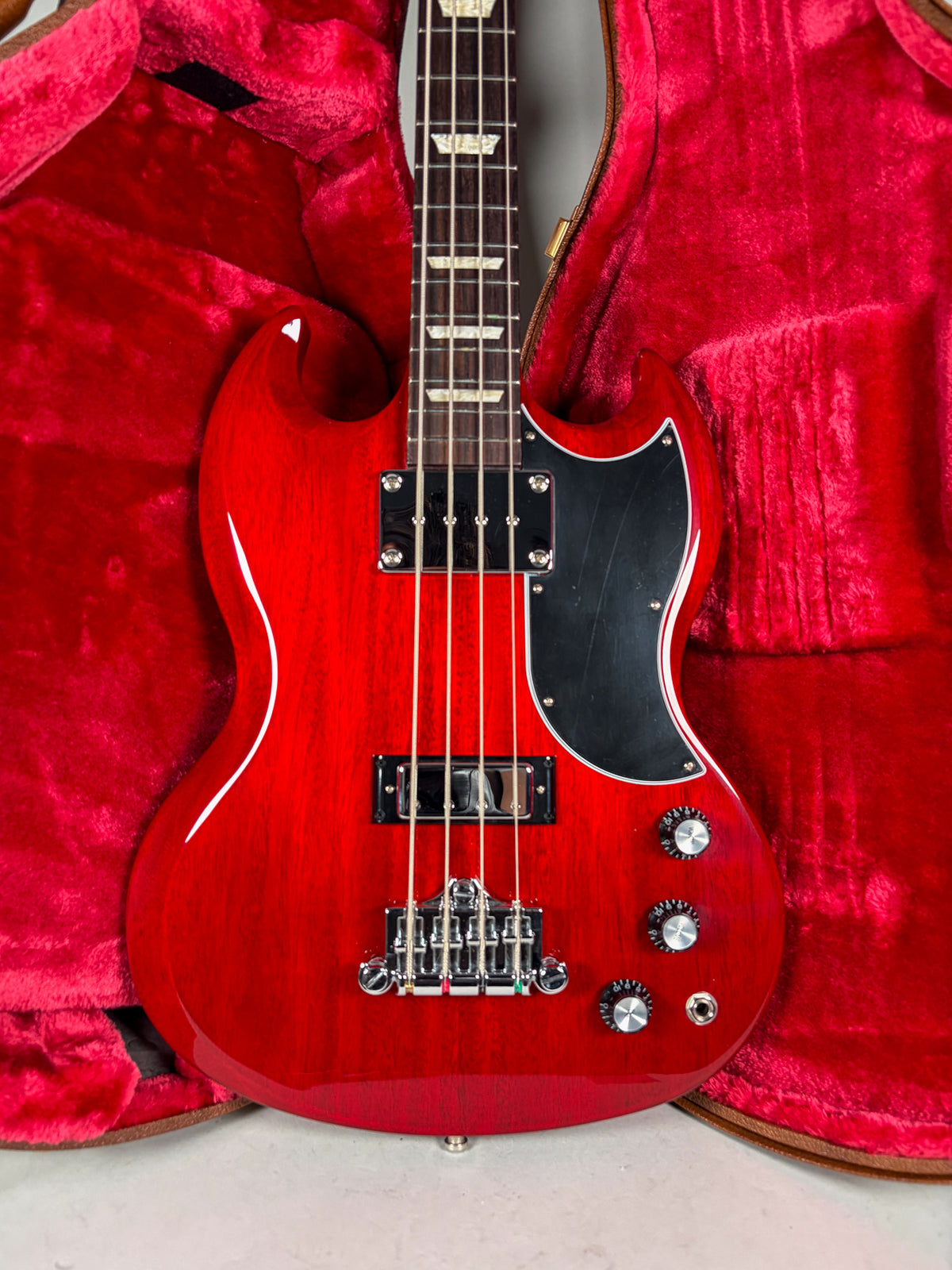 Full-front view of a cherry red bass guitar with a black pickguard, chrome hardware, and four black control knobs, resting in a red velvet case.