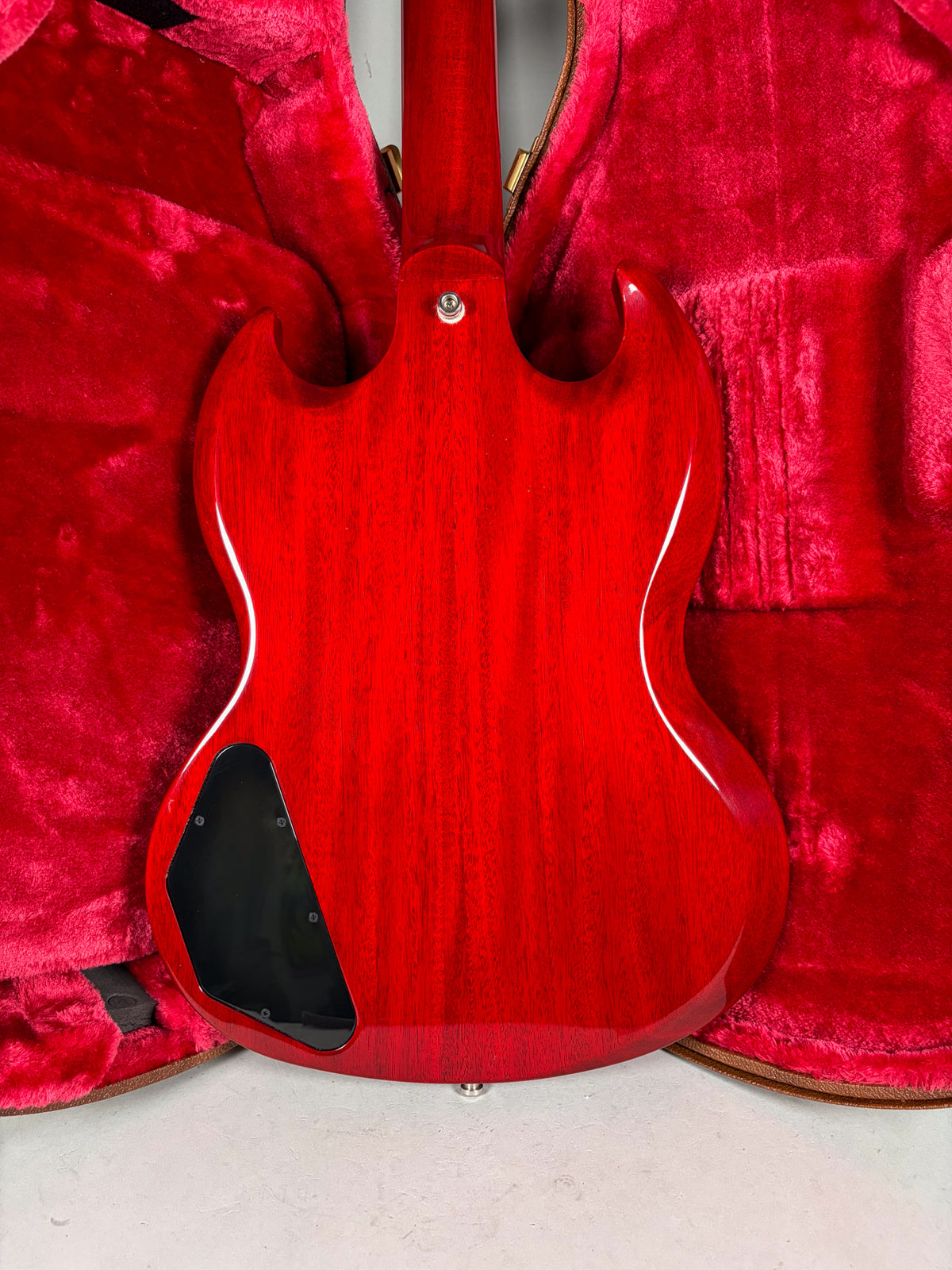 The back of a cherry red SG bass with a glossy finish, showing wood grain, a black control plate, and a neck plate.