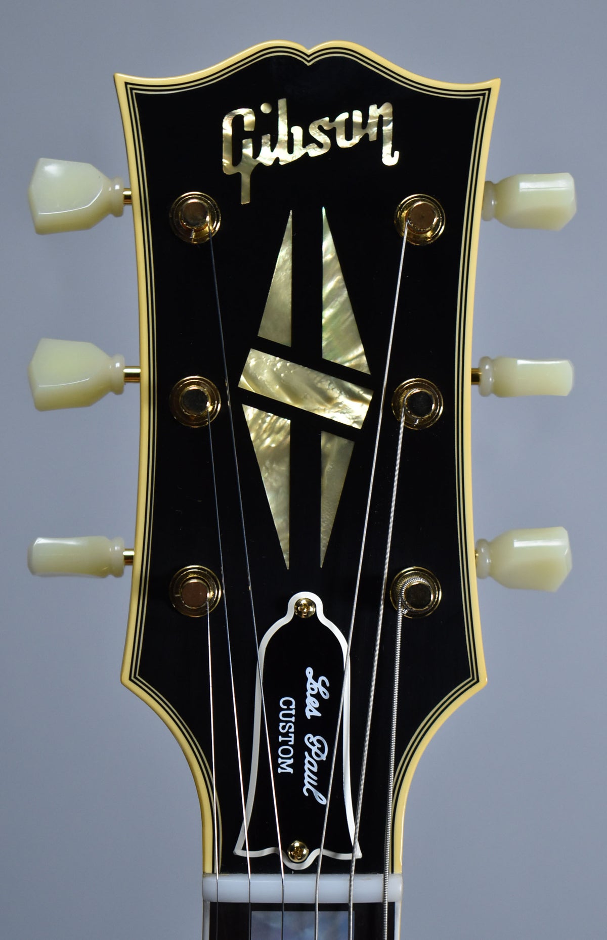 Close-up of a black headstock with cream binding, mother-of-pearl inlays, gold tuners, and a white truss rod cover.