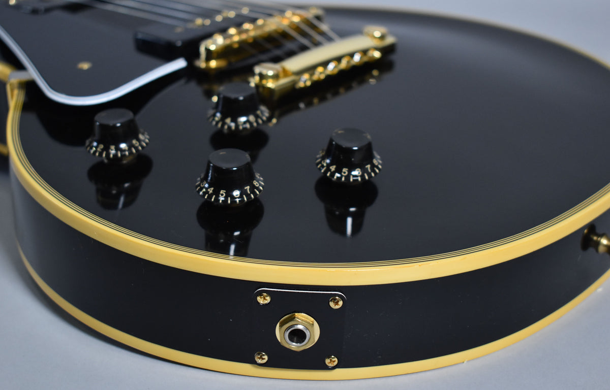 Close-up of a black electric guitar body with cream binding, gold hardware, black knobs, and an output jack.