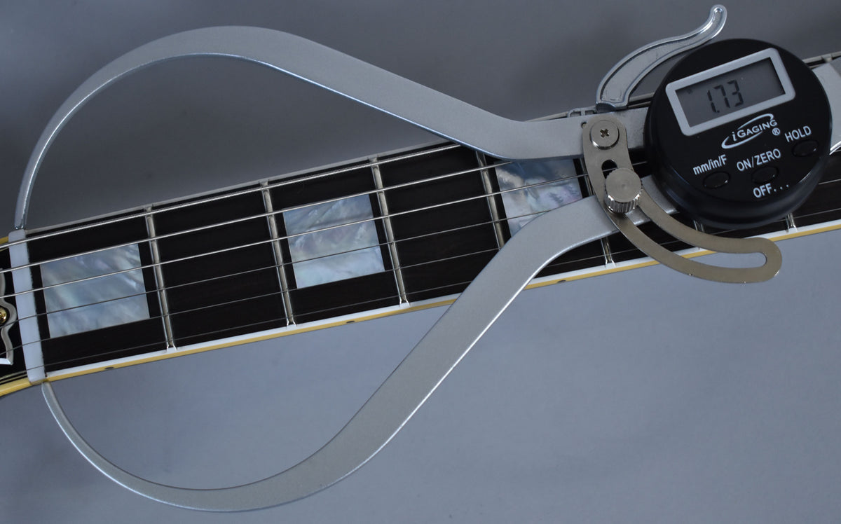 A digital caliper measures the guitar's fretboard, showing mother-of-pearl block inlays and a cream binding along the neck.