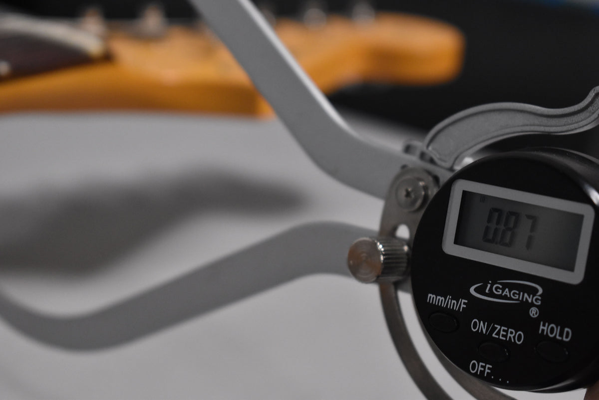 A digital caliper with a 0.87 reading is shown in the foreground, with a blurred guitar neck visible in the background.