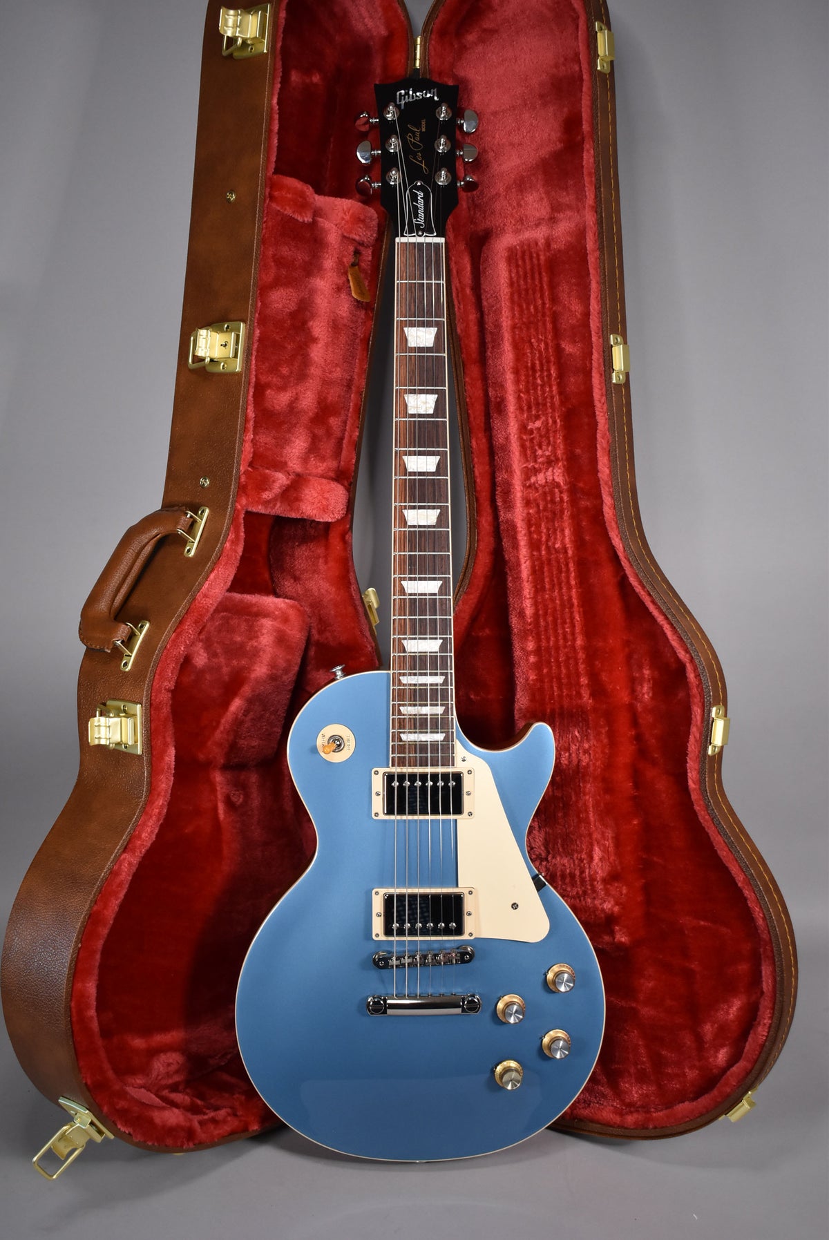 A Pelham Blue electric guitar with a cream pickguard and black humbuckers, resting in a brown hard case with red plush lining.