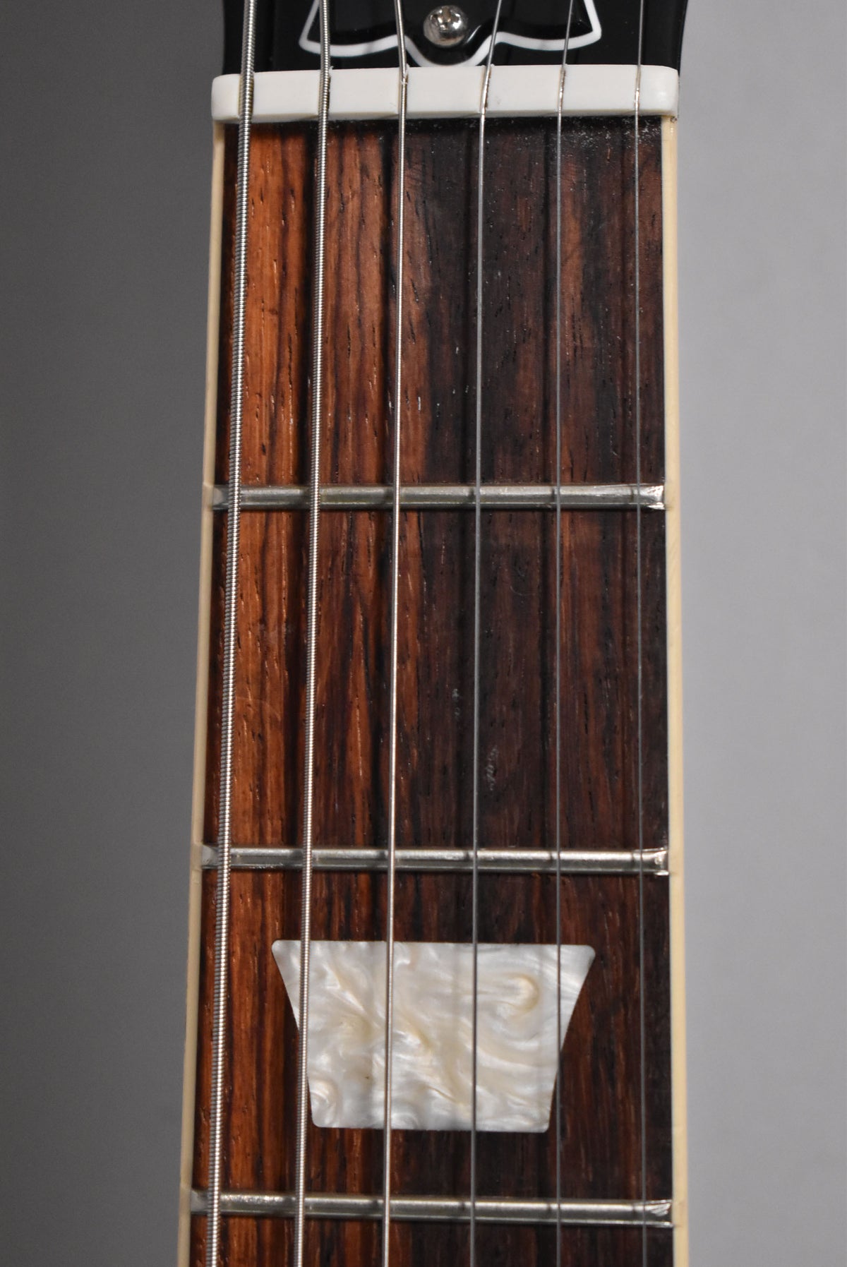 Close-up of the guitar's dark rosewood fretboard, showing the first five frets, a white nut, and a pearloid trapezoid inlay at the third fret.