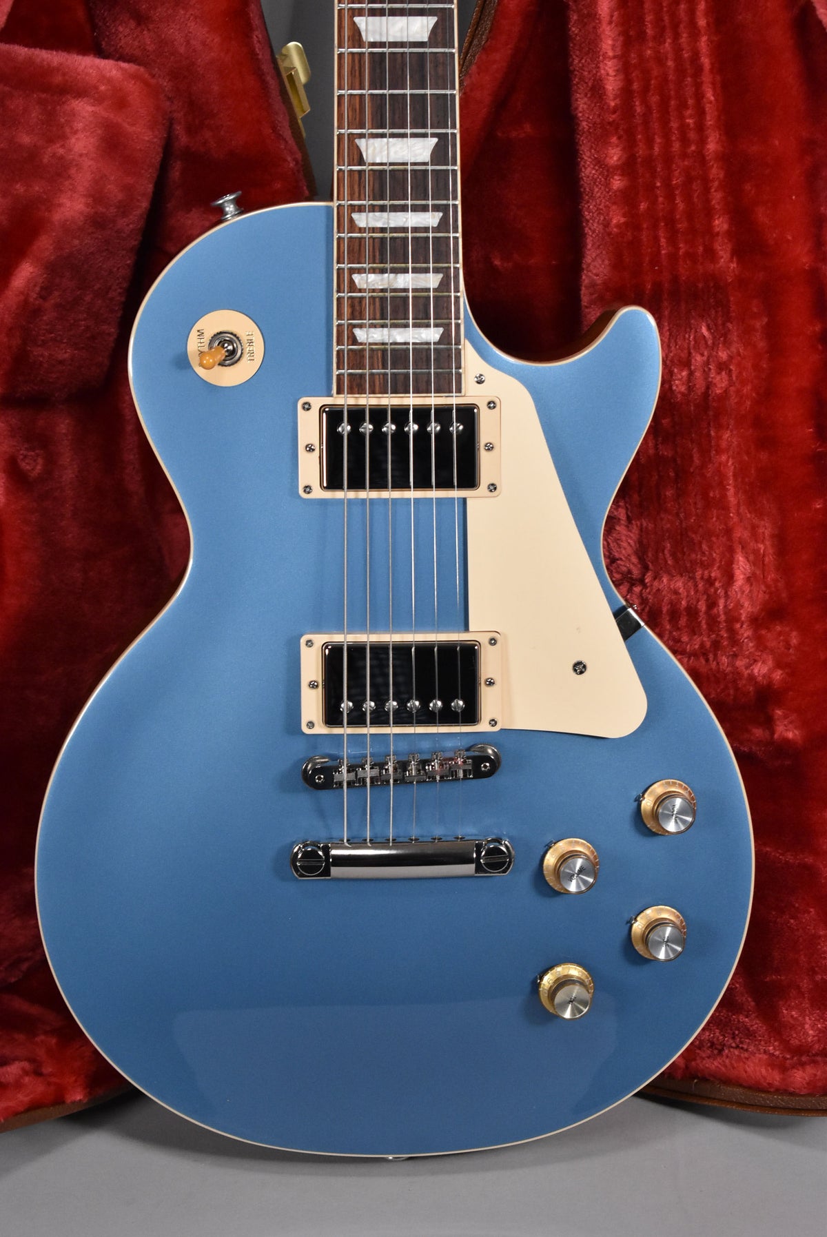 Pelham Blue Les Paul, body and neck visible, with cream pickguard, humbuckers, and gold top hat knobs, against a red case.