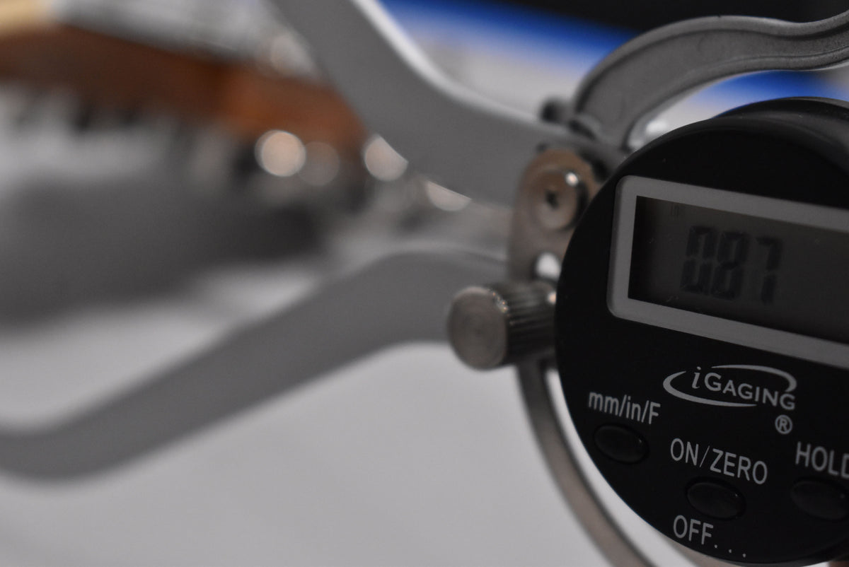 Close-up of a digital caliper showing a 0.087 measurement, with a blurred guitar neck and frets in the background.
