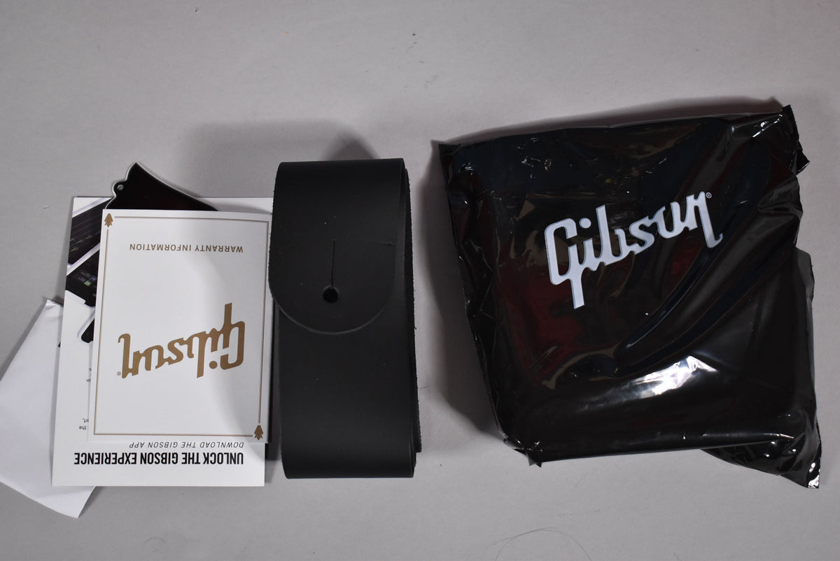 Overhead view of a black leather guitar strap, a warranty card, and a black plastic bag with a white logo.