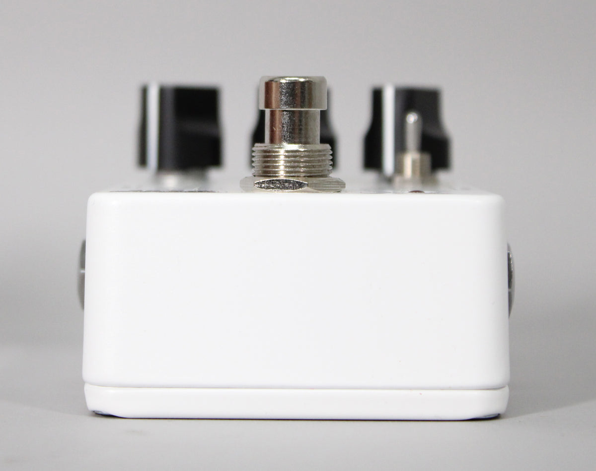 The white pedal is viewed from the front, showing a silver footswitch and two black control knobs on top.