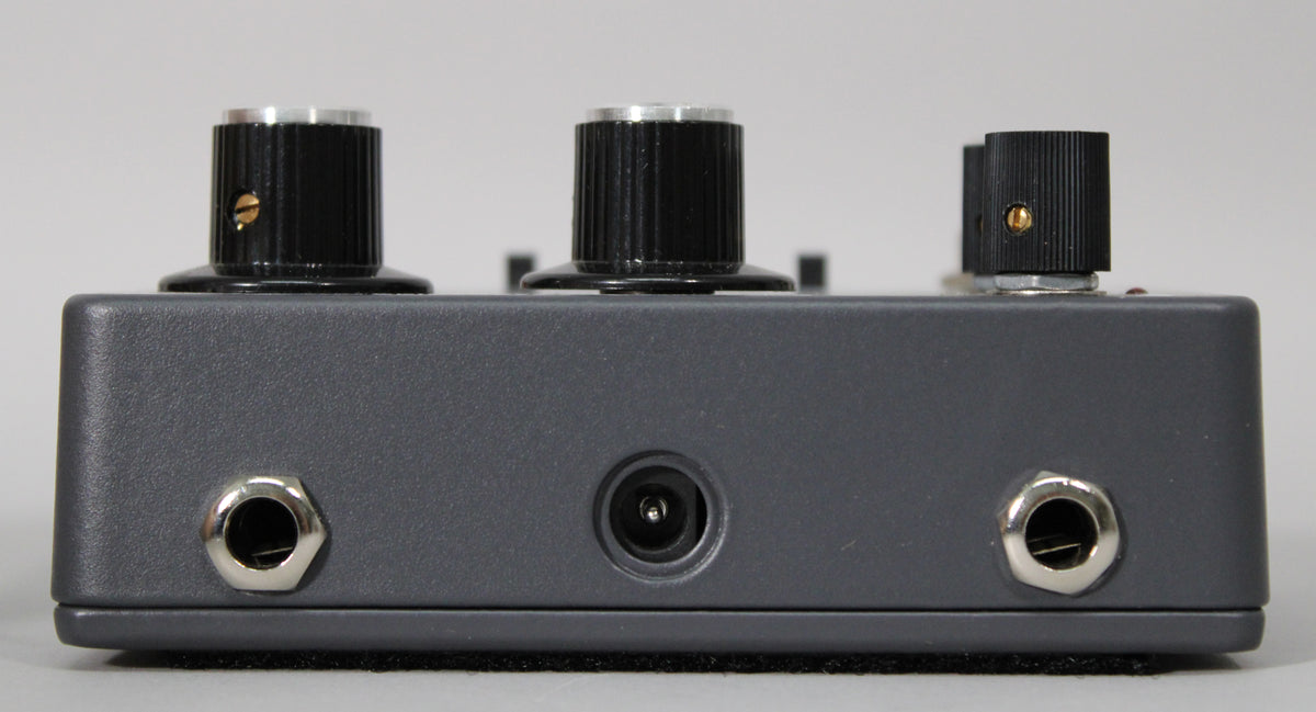 The dark grey pedal's rear shows two silver jack inputs, a power input, and three black fluted knobs with silver tops.
