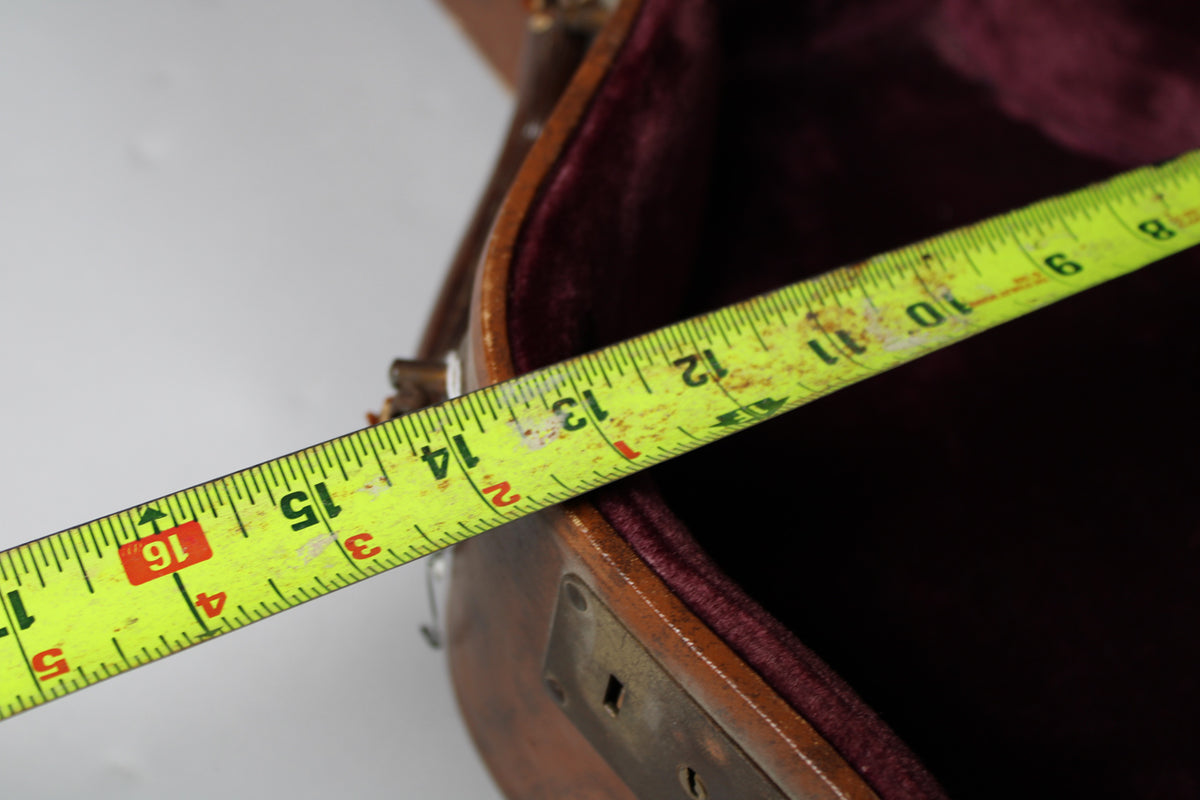 A yellow tape measure stretches across the open brown case, showing the pink interior and a tarnished metal latch.
