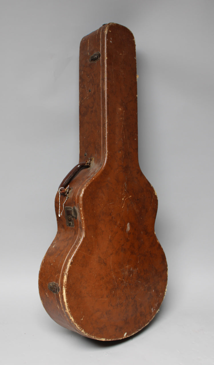 A brown archtop guitar case stands upright, showing its front and left side, with worn edges and scuffs on its textured surface.