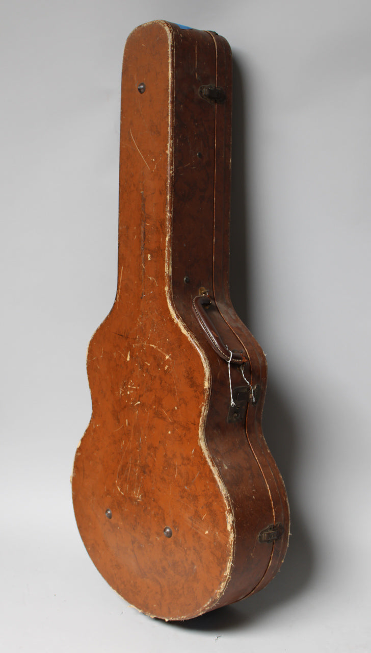 The brown archtop hardcase is shown upright, revealing its worn, scuffed exterior, leather handle, and two metal latches.