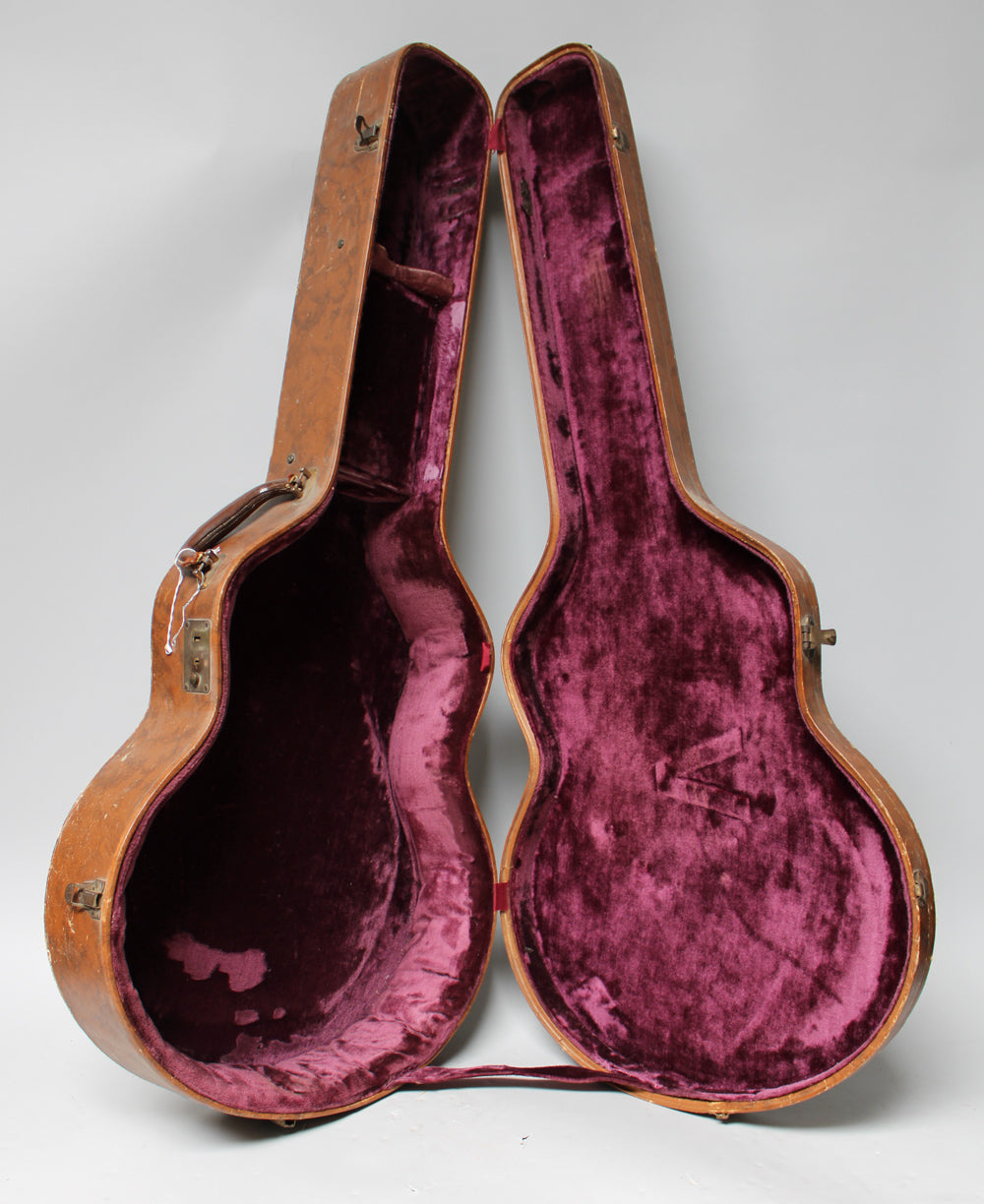 The open brown archtop case stands upright, revealing its worn pink velvet interior with a small accessory compartment.
