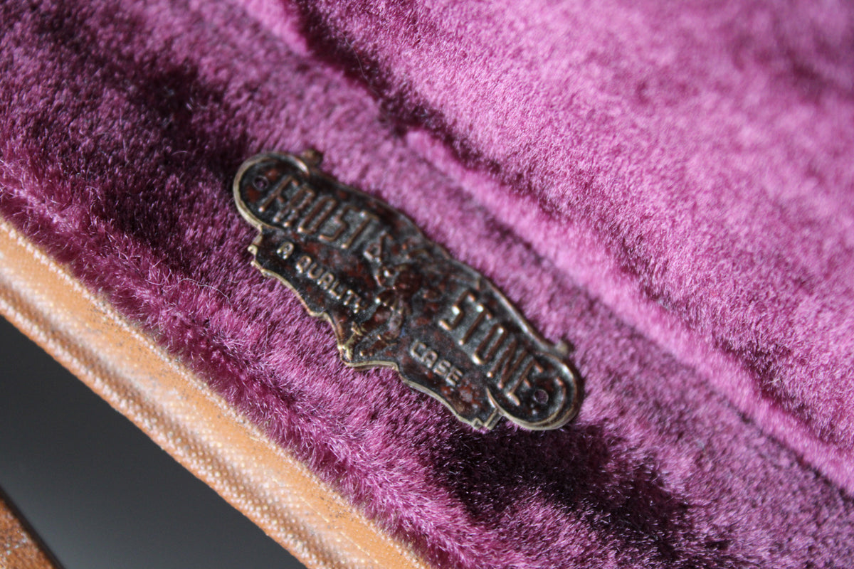 Close-up of the case's pink plush interior, showing a tarnished metal badge with raised lettering and a worn, light brown binding.