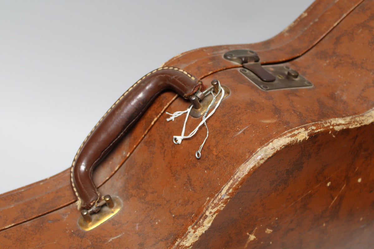 Close-up of the worn brown archtop case, showing the dark brown leather handle, a brass latch, and white string tied to a pin.