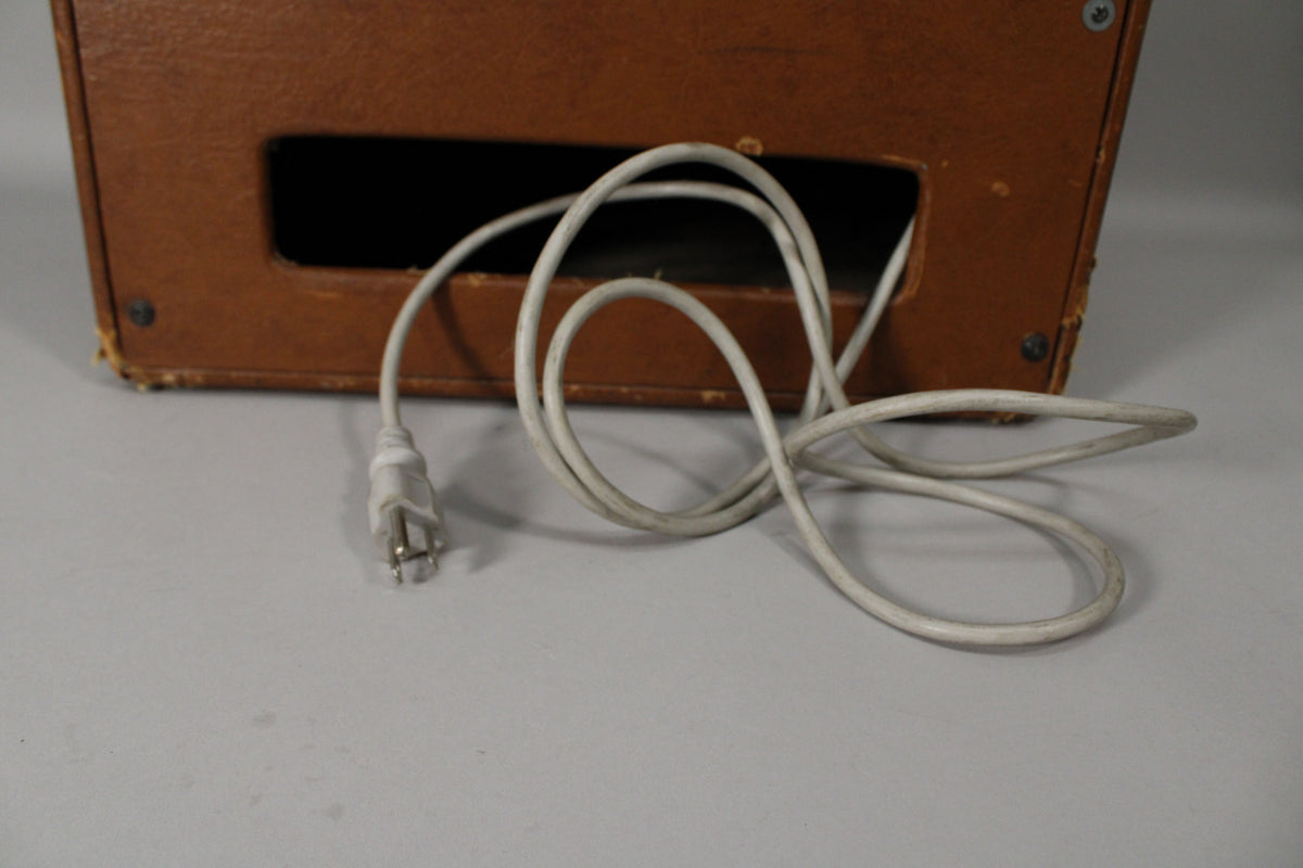 The amp's lower back panel, in worn brown tolex with a rectangular cutout, shows a coiled white power cord with a two-prong plug.