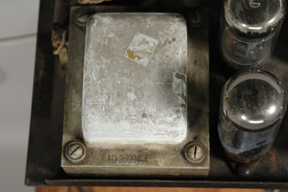 Overhead view of the amp's chassis, showing a rectangular, weathered silver transformer with a worn label, and two glass vacuum tubes.