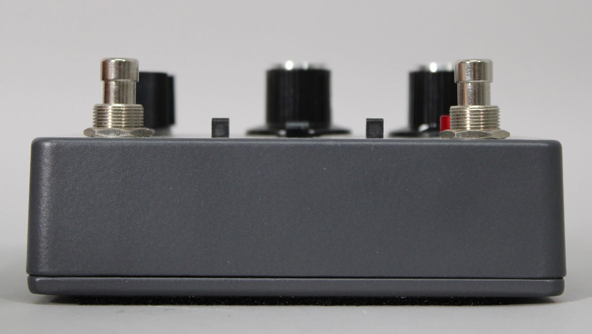 The dark gray pedal is viewed from the side, showing two silver jacks, two black knobs, and two small black feet.