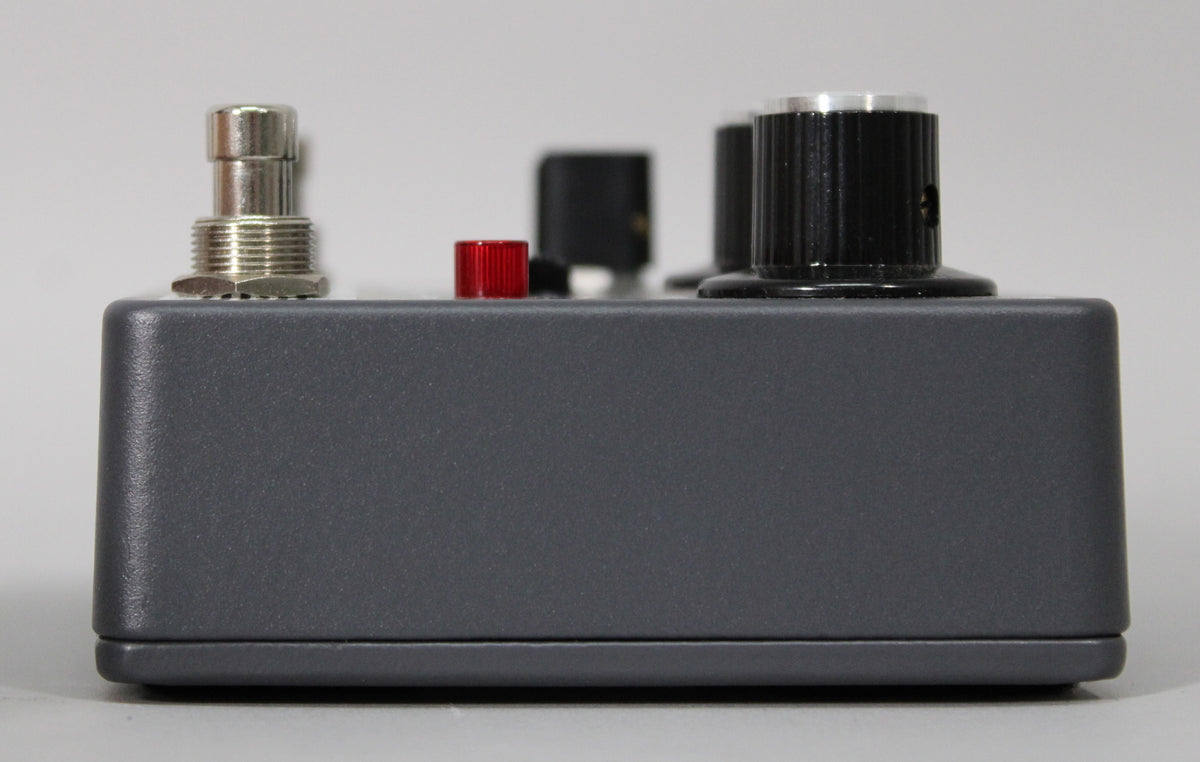 Side view of the dark gray pedal, showing a silver footswitch, a red button, and two black ridged knobs with silver tops.
