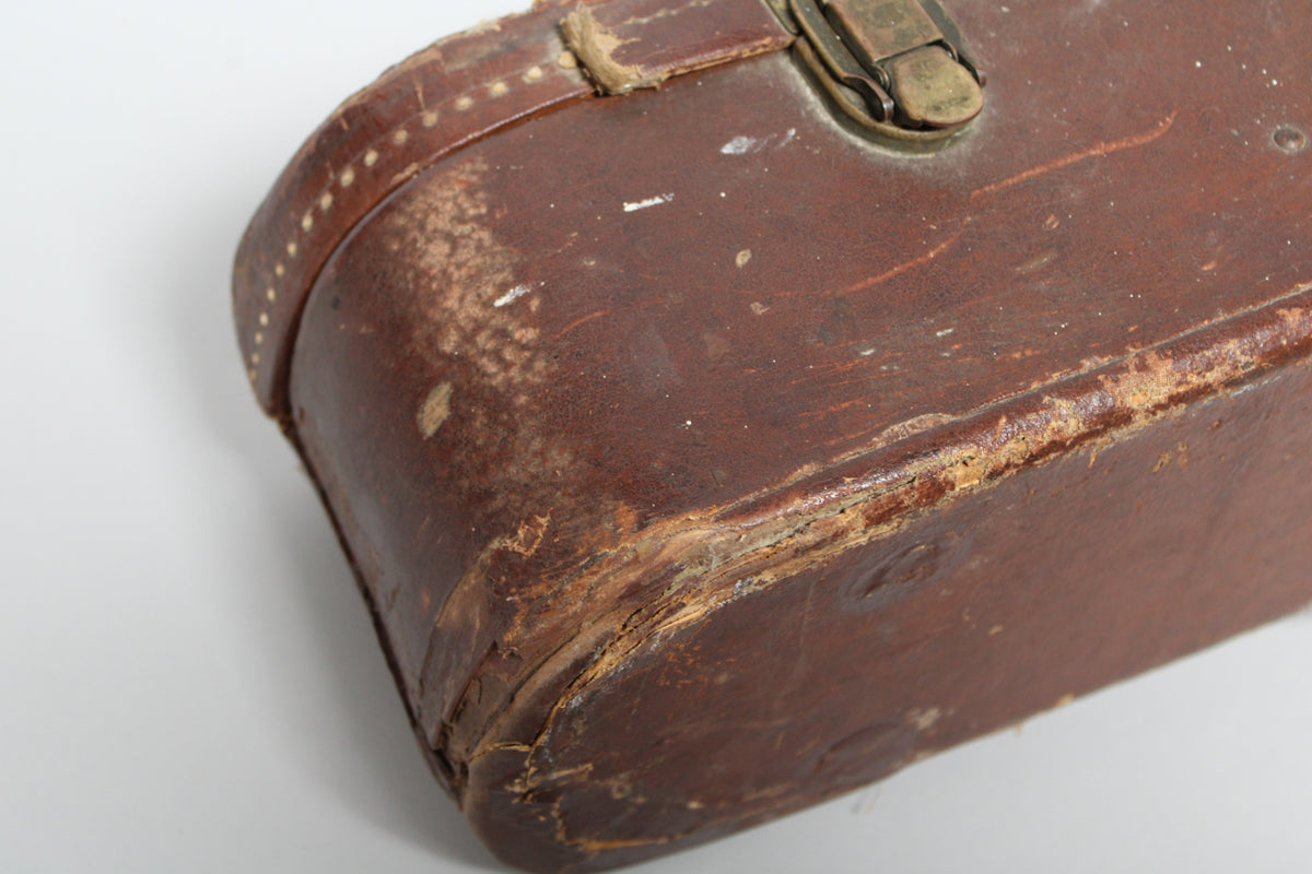 Close-up of a worn brown guitar case, showing a brass latch, scuffs, and exposed wood on the edges and corners.