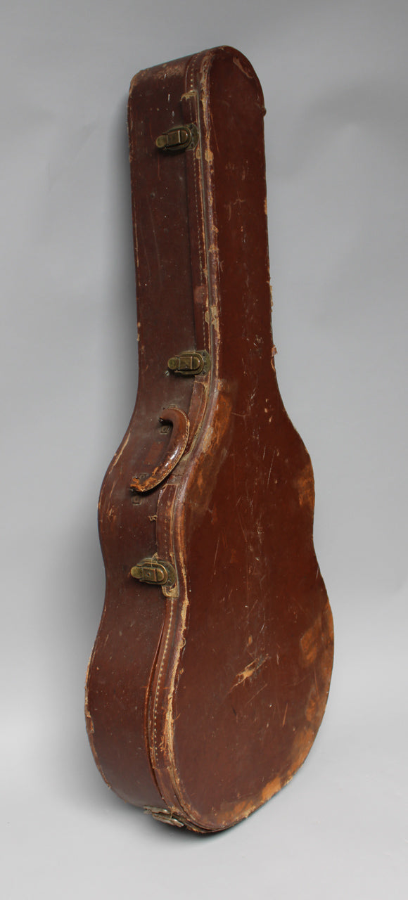 A brown guitar case stands upright, showing its side with a worn handle, three brass latches, and scuffed, peeling edges.