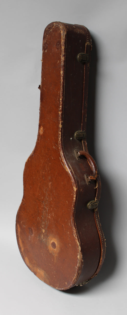 The brown guitar case stands upright, showing its worn, scuffed finish, a dark brown handle, and two tarnished latches.