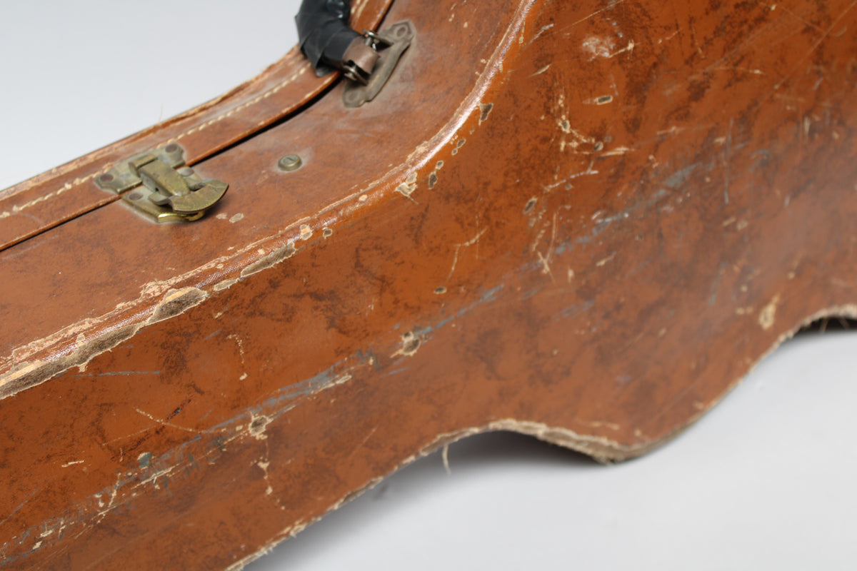 Close-up of a worn brown guitar case, showing a brass latch, black handle, and extensive scuffs and peeling on the surface.
