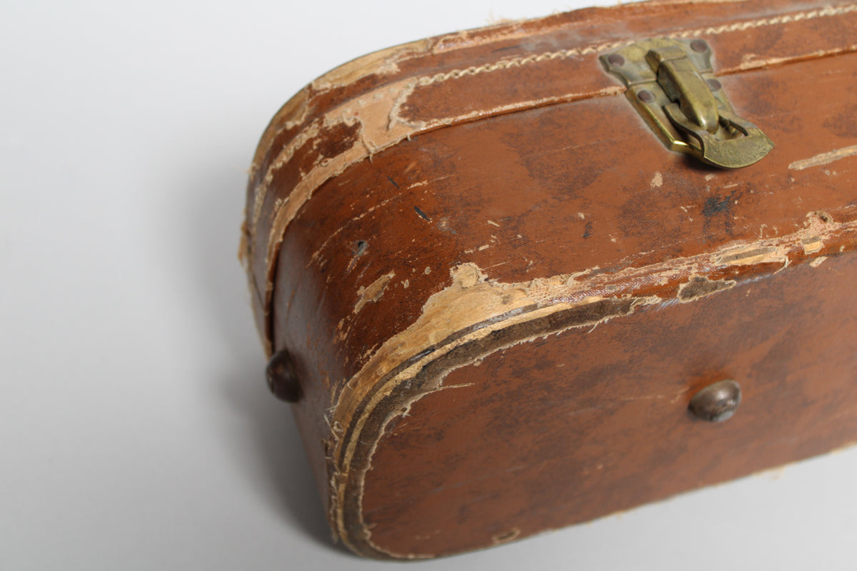 Close-up of a worn brown guitar case, showing peeling tolex, a brass latch, and two metal feet on the bottom edge.