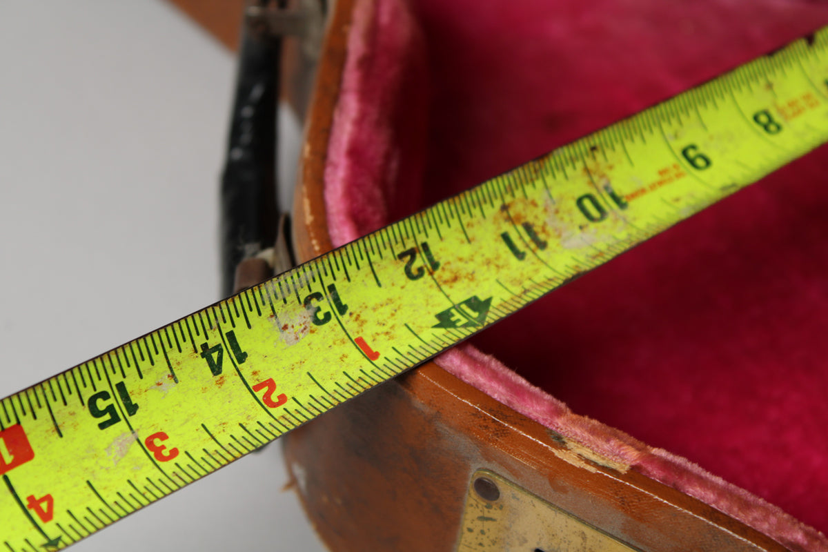 A yellow tape measure extends across the open brown case, showing measurements over the pink velvet interior.