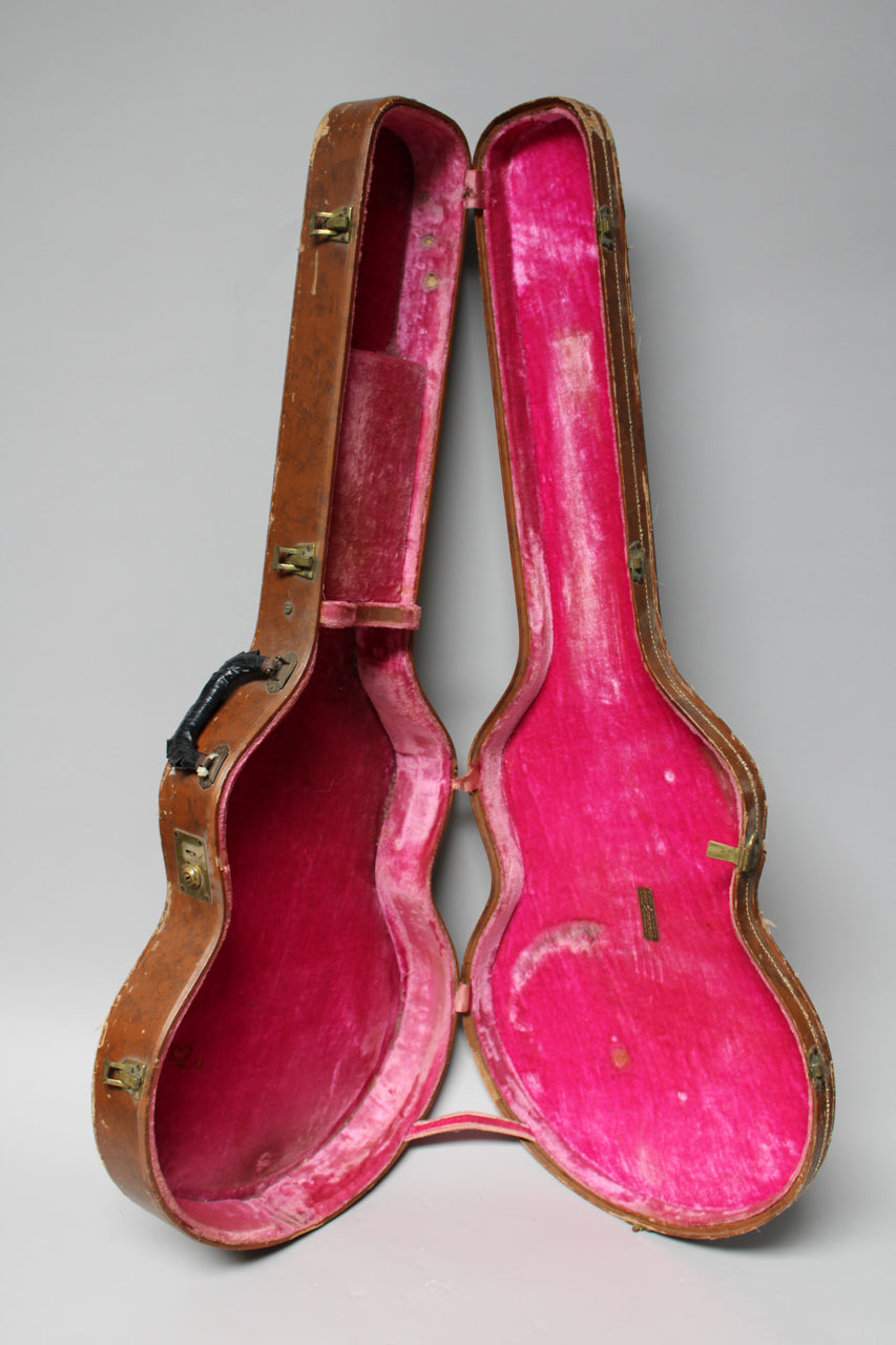 Open brown guitar case with worn pink velvet interior, showing a black handle and brass latches on the left half.