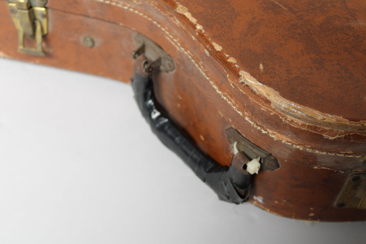 Close-up of the brown case's handle, wrapped in black tape, with significant wear and exposed wood around the edges.