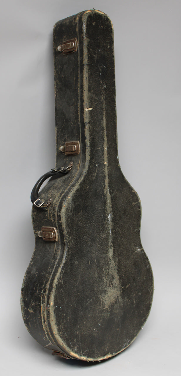 A black archtop guitar case stands upright, showing its worn, textured finish, three tarnished latches, and a black leather handle.
