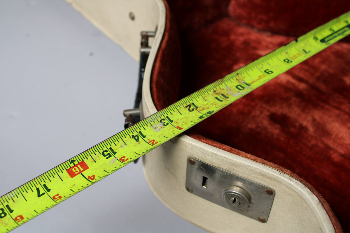 A yellow measuring tape stretches across the open tan case, showing the red velvet interior and a tarnished metal lock.